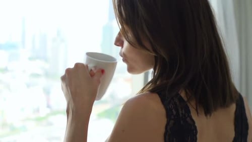 Woman drinks coffee by bright apartment window