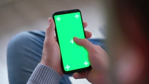 Young Man Sitting at Home Holding Smartphone Green Mockup Screen in Hand Male Person Using Chroma