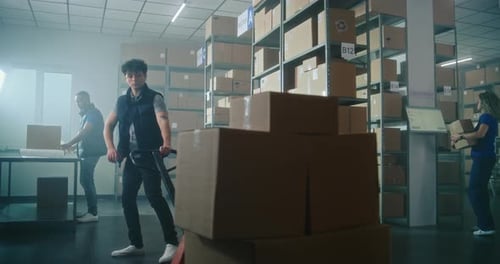 Male Inventory Specialist Pushes Pallet Truck with Cardboard Boxes in Warehouse Facility