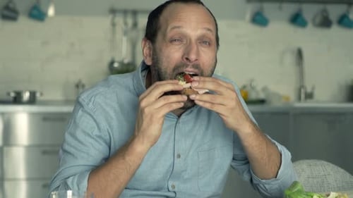 Happy, Young Man Eating Sandwich Sitting in Kitchen at Home 30s