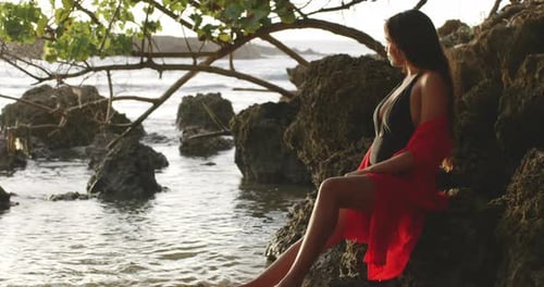 Latino Girl Model with Golden Tan Sitting on Tropical Beach Rocks at Sunrise Enjoys View, copy-space