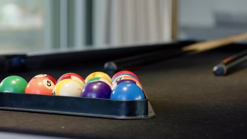 Camera is pulling back revealing racked billiard balls on black felt table near window showing cues