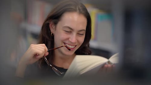 A Young Woman Sits in a Library Deeply Engrossed in a Book with a Thoughtful Expression