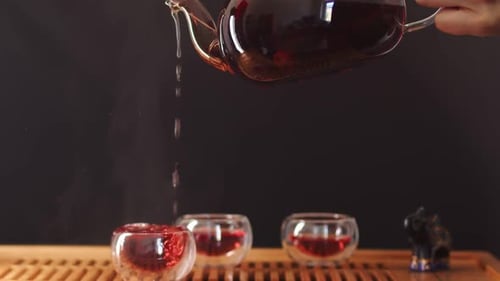 Klare Teetassenüberläufe mit heißem Teegetränk, das aus einer hoch darüber gehaltenen Glasteekanne fließt, zwei halbvoll