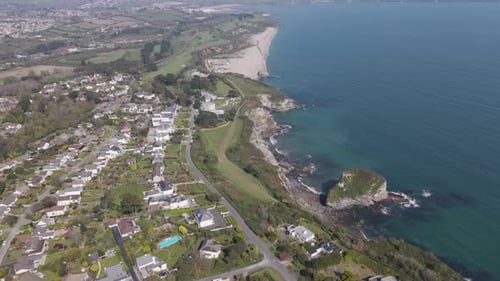 Aerial sweep over sandy arc of coastal bay where hillside homes meet verdant cliffs and shimmering s