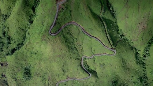 Drone footage of a winding mountain road cutting through lush green hills.
