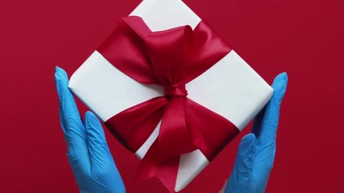 Medical Gloves Holding White Gift Box With Red Ribbon