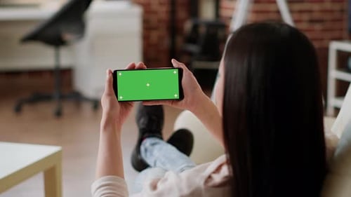 Woman Sitting on Sofa While Holding Smartphone with Green Screen Mockup Template Background