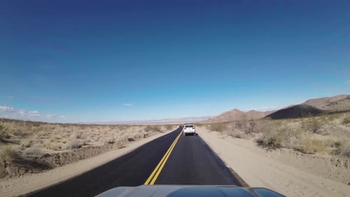 Ein Fahrzeug folgt einem anderen auf einer asphaltierten Wüstenstraße unter klarem blauem Himmel in der Nähe des Joshua Tree National