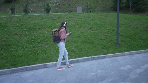 Young Woman Reading Book Walking Park Pathway Wearing Backpack and Headphones Enjoying Music From
