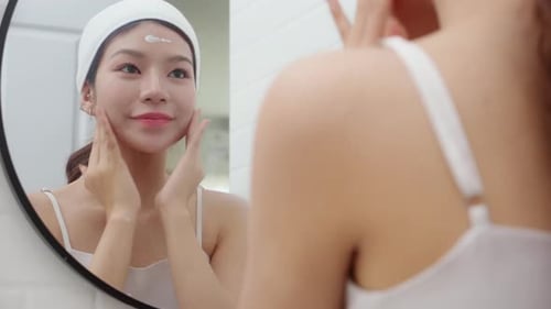Schöne junge Frau, die Creme oder Lotion zur Verjüngung der Gesichtshaut aufträgt und dabei in den Spiegel schaut.