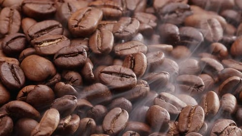 Close up coffee beans are roasted and rotated clockwise, there is a little smoke.