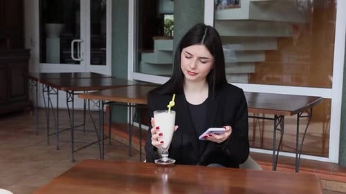 91A girl uses a smartphone and drinks a cocktail while sitting at a table on the street.