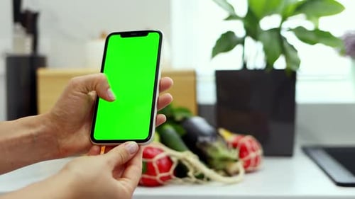 Close-up of female hands holding smartphone with green screen on a background of organic vegetables