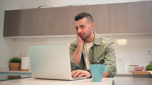 Tired Man Working on Laptop in Kitchen