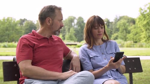 An Unhappy Middleaged Man Tries to Talk to a Middleaged Woman Who Works on a Smartphone in a Park