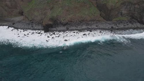 Luftaufnahme von Wellen, die in einen Kieselstrand einschlagen, türkisfarbenes Wasser und weißer Schaum. Drohne bewegt sich nach links