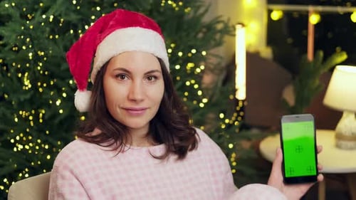 A Woman in a Christmas Cap Holds Up a Smartphone with a Green Screen in Her Hand