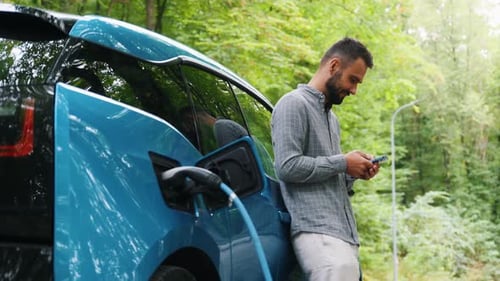 Man stands with phone near her electric car and waits when vehicle will charged at green forest. Sav