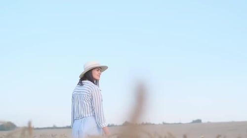 A Young American Woman in a Hat Walks Through a Wheat Field Against the Background of Calm Skyblue