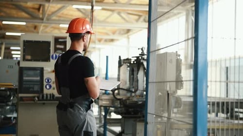 Manager in orange hard hat stands in the factory with machines and writes information in notepad