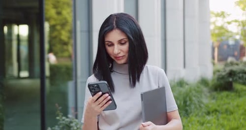 Young caucasian business woman using cell phone mobile app and holding computer device for work, tra