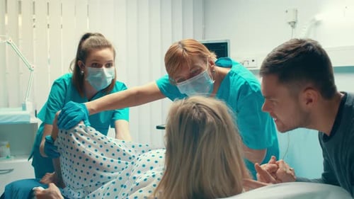 En el hospital, una mujer que da a luz, su esposo la sostiene de la mano en señal de apoyo y los obstetras la ayudan. Modificar