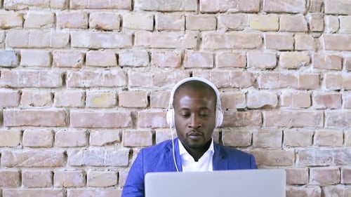 Man with headphones working on laptop by brick wall