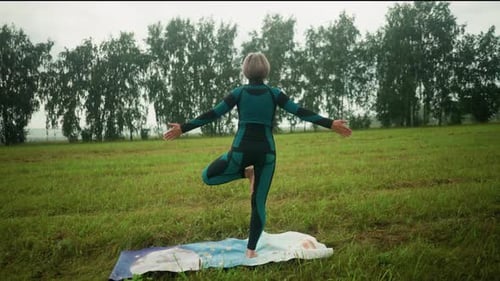 Back View of Woman in Tree Pose Practicing Yoga in a Grassy Field with Distant Trees