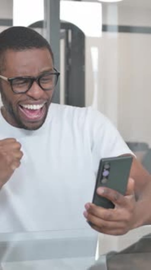 Young African Man Celebrating Success on Phone in Office, vertical video