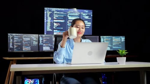 Une programmeuse asiatique boit du café tout en écrivant du code sur un ordinateur portable à l'aide de plusieurs moniteurs