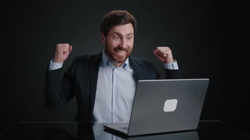 Ein Mann in einem schwarzen Anzug, der etwas auf dem Laptop tippt und dann jubelt, als Geld auf ihn fällt
