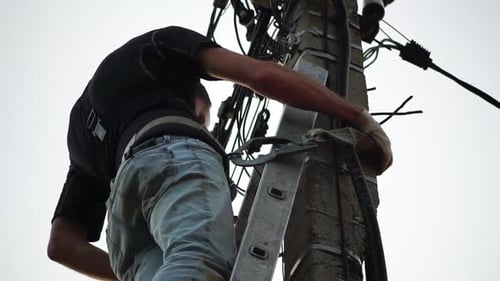 Skilled Electrician Fixes Wires Standing on Ladder Near High Pole Against Blue Sky on Summer Day
