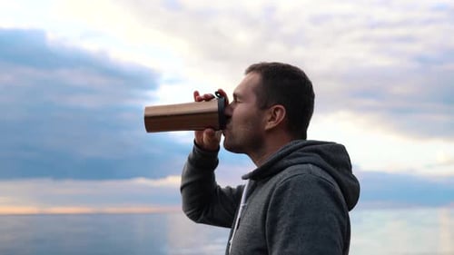 A Man Drinking Coffee or Tea From a Warm Thermos Against the Backdrop of a Sunset in a Calm Sea or