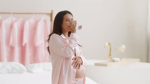 Junge glückliche schwangere Frau im Pyjama trinkt Wasser und streichelt ihren Bauch und genießt einen gesunden Start