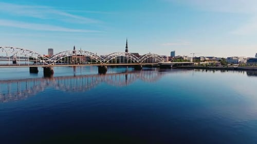 Drone Flies Toward Riga Railway Bridge and Skyline – Calm Morning Reflection