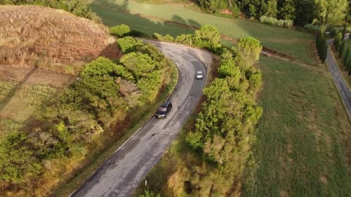 Drone filmé en train de suivre une voiture blanche sur la route qui contourne une colline pendant qu'une autre voiture passe.
T