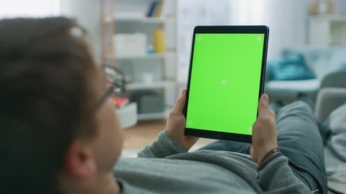 Young Man at Home Laying on a Couch Using with Green Mock-up Screen Tablet Computer in Vertical Por