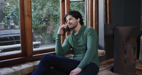 Happy caucasian man talking on smartphone sitting by window at home, slow motion