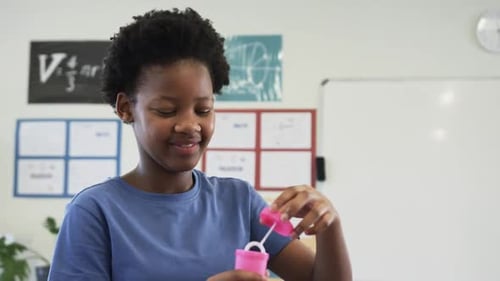 African teen female unscrewing pink bubble bottle dipping wand blowing bubbles in classroom for fun