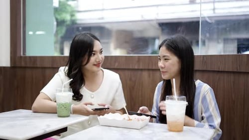 Two young asian women friends sitting at coffee shop having a coffee and chatting with smiling laugh