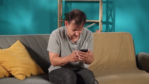 A Nervous Twitchy Man Sits on a Living Room Couch Using a Smartphone and Playing an Online Game The