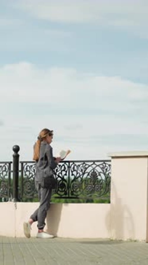 Lady in Grey Reading By Fence with Trees and Lamp Post in Background