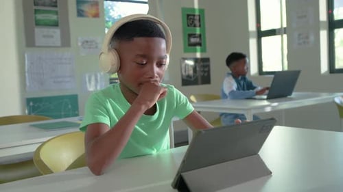 African teenage boy wearing beige headphones tapping tablet folio, following lesson at school desk