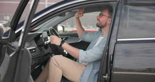 Young Caucasian Man Sits in a New Car on a Outdoor Parking Lot and Inspecting Interior
