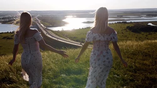 Two Young Happy Blonde Women in Light Summer Dresses Holding Hands and Walks to the Edge of the Hill