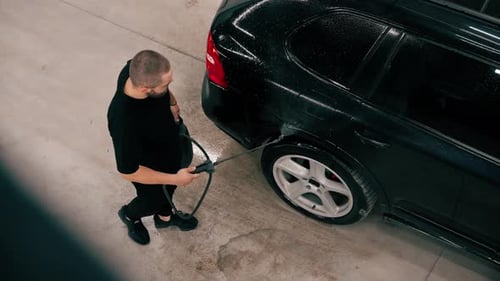 A male car wash employee applies car wash detergent to a black luxury car using spray gun in the car