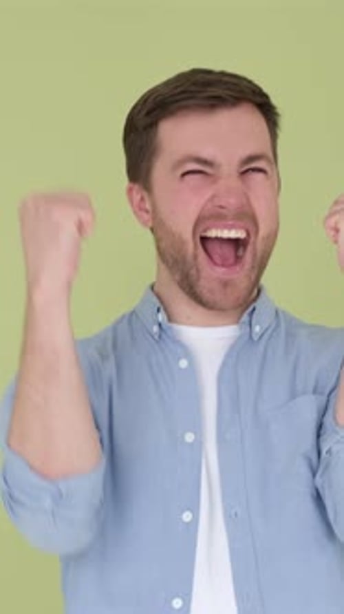 Emotionaler junger Mann hebt beiläufig die Fäuste auf dem Hintergrund von Olive Studio und feiert Erfolg Vertikal