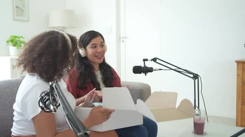 Female cohosts opening box on couch with boom mic, revealing white LED mask during podcast