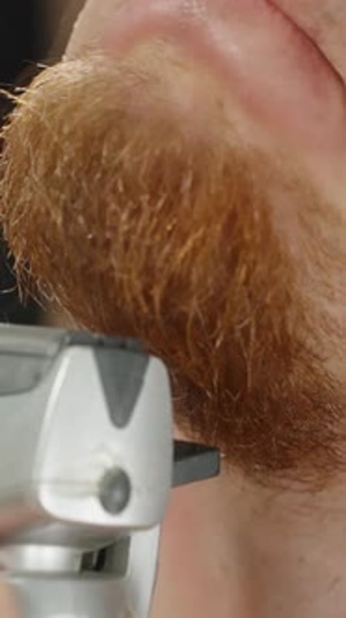 Vertical video. Man with Red Beard Trimming It with Electric Trimmer, Close-Up in Front of Mirror.